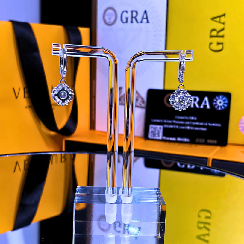 Elegant flower-shaped moissanite drops with sparkling halo setting on huggie hoops in sterling silver. Beautiful movement and brilliant fire enhance any look effortlessly.