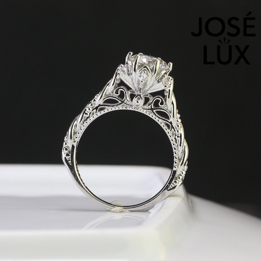 Intricate filigree detailing and ornate vintage craftsmanship create a timeless design featuring brilliant round moissanite with delicate metalwork accents.