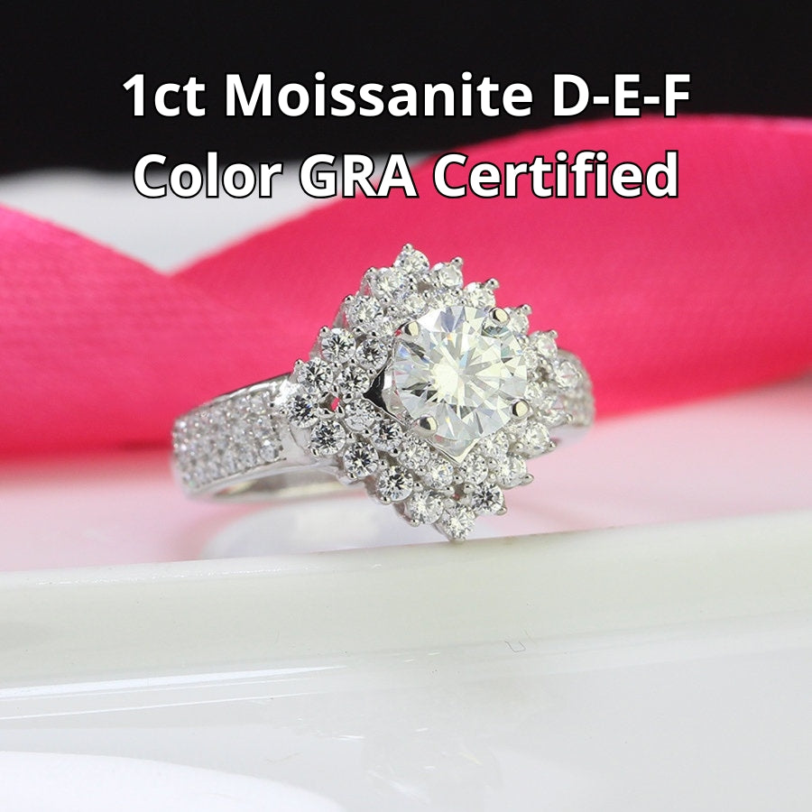 Brilliant round moissanite centerpiece enhanced by dual layers of sparkling accent stones with pavé detailing on the polished silver band.