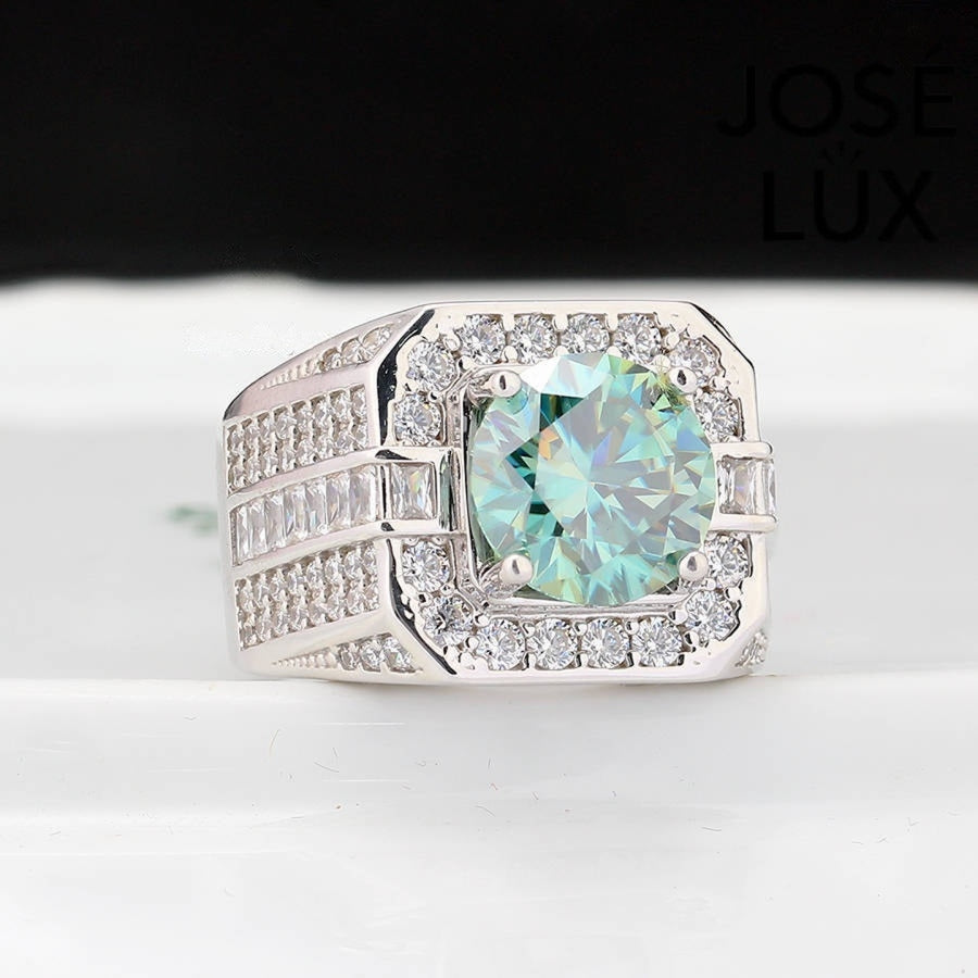Substantial sterling silver men's ring featuring a 4-carat round green moissanite encircled by a cushion-shaped halo. Wide band with alternating rows of round and baguette stones creates impressive depth and dimension.