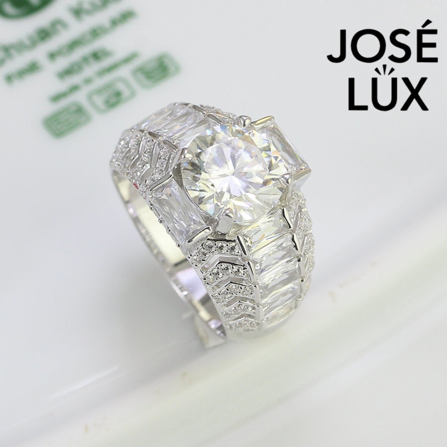 Commanding men's ring in sterling silver features a brilliant 4-carat round moissanite in six-prong setting. Wide band showcases graduated baguette-cut stones in chevron pattern with pavé accents for dimensional impact.